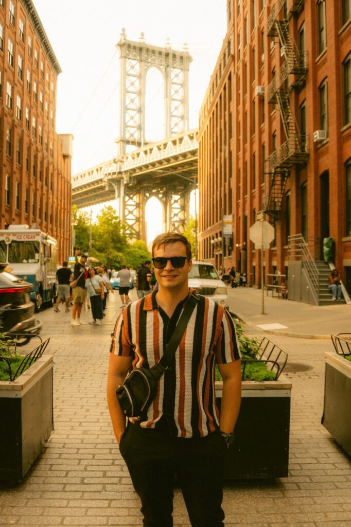 Kyle smiling on a cobblestone street in Dumbo, Brooklyn, with the Manhattan Bridge behind him