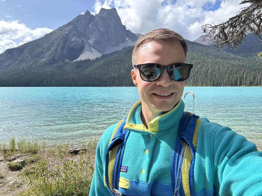 Kyle smiling in a teal fleece and sunglasses in front of a mountain lake